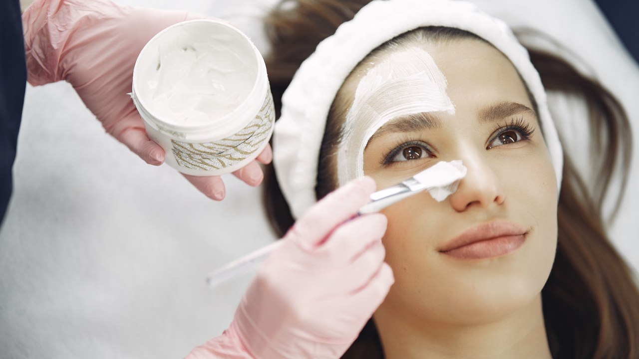 Professionista applica una maschera cosmetica bianca con un pennello sul viso di una donna durante un trattamento estetico.